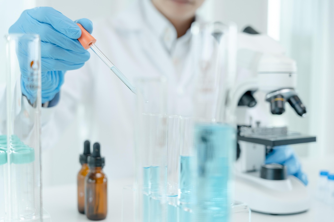 A person wearing a white lab coat and latex gloves holds a dropper of blue liquid above a test tube of the same fluid. There are other test tubes and a microscope on the table in front of the person.